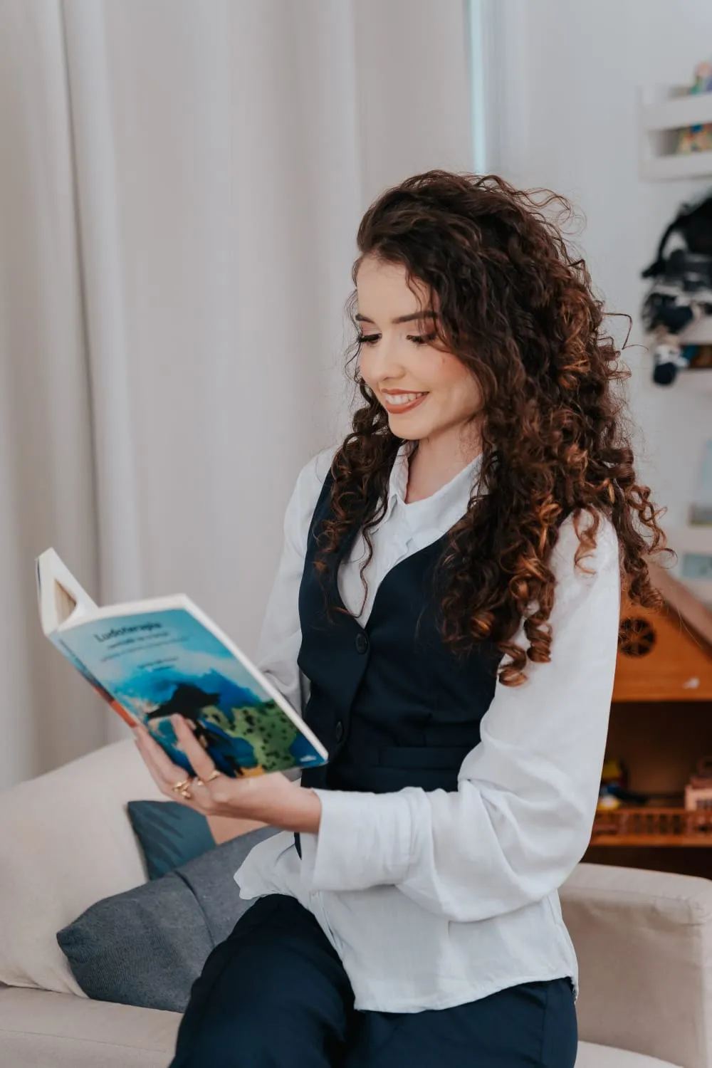 Alice Lins sorrindo enquanto lê um livro, sentada em um sofá claro.