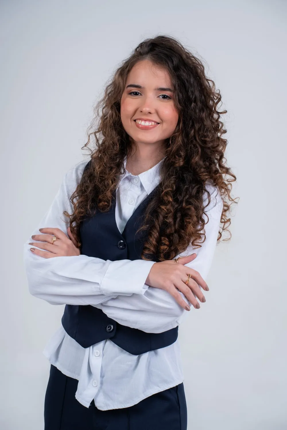 Retrato de Alice Lins, psicóloga: sorridente, de cabelos cacheados longos, de colete azul-marinho e camisa branca, com os braços cruzados.